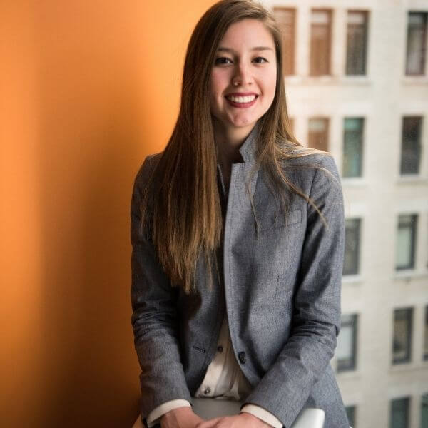 Girl holding a folder sitting in a formal environment. She is wearing a shirt, a blazer and a pair of formal pants.